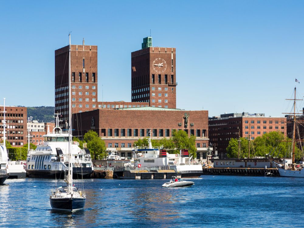 A sail boat sails from the marina in the Oslo fjord in front of the City hall building in Norway capital city on a sunny day.